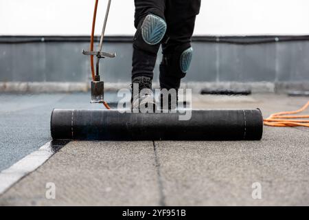 Professioneller Dachdecker, der Bitumenwalze auf Flachdach mit einem Gasbrenner auf einer modernen Baustelle aufbringt Stockfoto