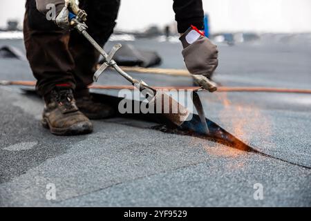 Professioneller Dachdecker, der Bitumenwalze auf Flachdach mit einem Gasbrenner auf einer modernen Baustelle aufbringt Stockfoto