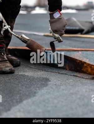 Professioneller Dachdecker, der Bitumenwalze auf Flachdach mit einem Gasbrenner auf einer modernen Baustelle aufbringt Stockfoto