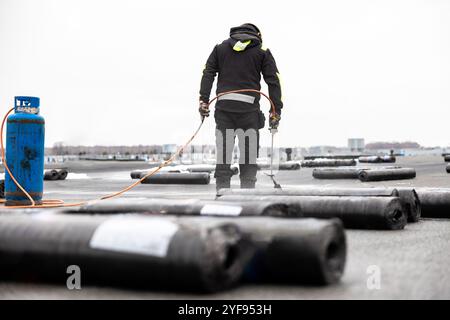 Professioneller Dachdecker, der Bitumenwalze auf Flachdach mit einem Gasbrenner auf einer modernen Baustelle aufbringt Stockfoto