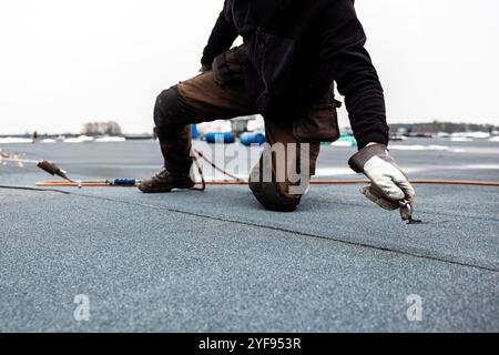 Professioneller Dachdecker, der Bitumenwalze auf Flachdach mit einem Gasbrenner auf einer modernen Baustelle aufbringt Stockfoto