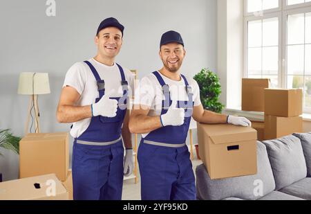 Zwei glückliche Arbeiter von der Transportfirma, die in einem Raum voller Kisten standen und die Daumen nach oben zeigen Stockfoto