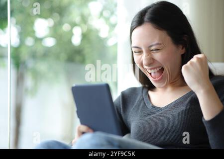 Aufgeregte asiatische Frau, die eBook liest, zu Hause sitzt Stockfoto