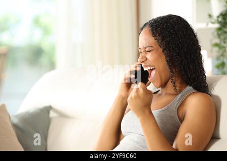 Aufgeregte schwarze Frau, die gute Nachrichten feiert und zu Hause telefoniert Stockfoto