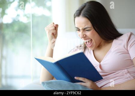 Eine aufgeregte Frau liest ein Papierbuch auf einer Sitzcouch zu Hause Stockfoto