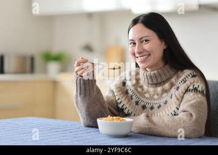 Glückliche asiatische Frau im Winter isst Müsli und schaut dich in der Küche zu Hause an Stockfoto