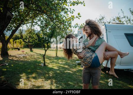 Ein verliebter Mann, der seine Verlobte in Waffen hält. Das junge Paar ist auf einem Campingausflug in die Natur unterwegs mit dem Wohnwagen. Stockfoto