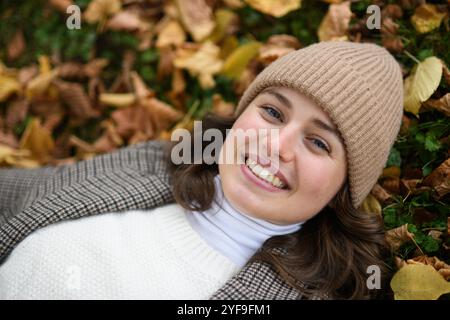 Porträt einer schönen Frau, die in herabfallenden Herbstblättern auf dem Rasen liegt. Sie lächelt und sieht in die Kamera. Stockfoto