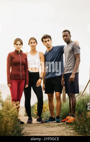 Vier Freunde stehen gemeinsam in sportlicher Kleidung auf einem malerischen Wanderweg im Freien. Sie strahlen Selbstvertrauen und Kameradschaft aus und sind bereit für ein Workout oder einen Lauf. Stockfoto