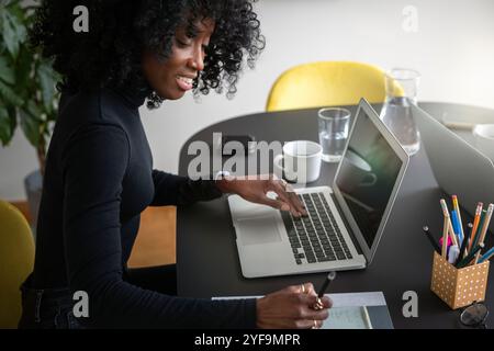 Lächelnde Geschäftsfrau, die auf Notizblock schreibt, während sie mit Laptop im Büro sitzt Stockfoto