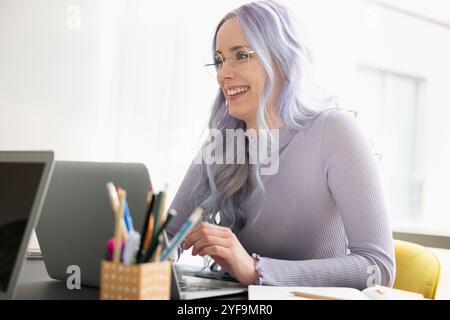 Glückliche Geschäftsfrau mit lila Haaren, die eine Brille trägt und mit Laptop im Büro sitzt Stockfoto