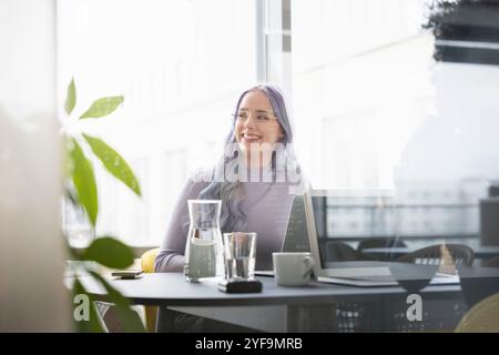 Glückliche Geschäftsfrau mit lila Haaren, die eine Brille trägt und im Büro wegblickt Stockfoto