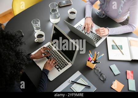 Hochwinkelansicht von Unternehmerinnen, die am Laptop arbeiten, während sie im Büro neben dem Tisch sitzen Stockfoto