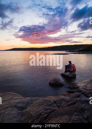 Blick auf einen Mann, der auf Felsen im Meer sitzt und den Blick während des Sonnenuntergangs betrachtet Stockfoto