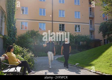 Glückliche männliche Freunde, die auf einem Fußweg laufen, begrüßen weibliche Nachbarin, die auf einer Bank im Garten sitzt Stockfoto