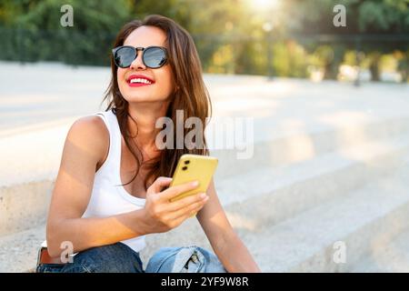 Urbane Frau in lässigem Outfit, die auf der Treppe sitzt und lächelt, während sie ihr Smartphone benutzt. Stockfoto