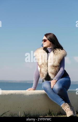 Eine stilvolle und modische Frau in einer Pelzweste sitzt auf einem Pier vor dem Hintergrund des Meeres Stockfoto