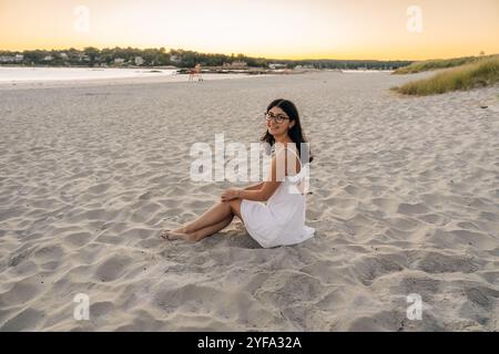 Frau sitzt am Sandstrand bei Sonnenuntergang und lächelt in einem weißen Kleid Stockfoto