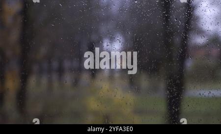 Ein Blick durch ein regendurchflutetes Fenster. Regentropfen zieren eine Fensterscheibe auf einem unscharfen Hintergrund mit der Silhouette von Bäumen in einem Park vor einer Stadt Stockfoto