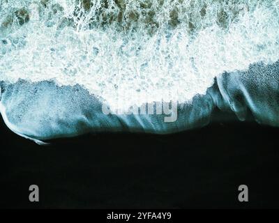 Aus der Vogelperspektive auf Islands schwarzem Sandstrand stürzende Wellen, die einen beeindruckenden Kontrast zwischen dunklem Sand und weißem Schaum schaffen und die raue Powe einfangen Stockfoto
