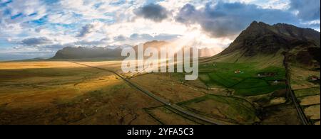 Atemberaubende Aussicht auf Islands majestätische Landschaft mit üppigen grünen Feldern, dramatischen Bergen und Sonnenstrahlen, die durch die Wolken dringen, und das zeigt sich Stockfoto