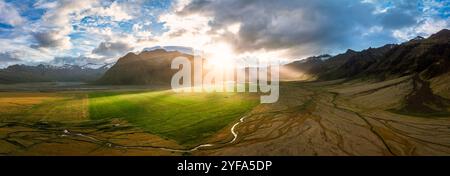 Atemberaubende Aussicht auf Islands majestätische Landschaft mit üppigen grünen Feldern, dramatischen Bergen und Sonnenstrahlen, die durch die Wolken dringen, und das zeigt sich Stockfoto