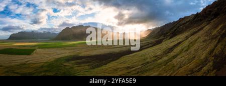 Atemberaubende Aussicht auf Islands majestätische Landschaft mit üppigen grünen Feldern, dramatischen Bergen und Sonnenstrahlen, die durch die Wolken dringen, und das zeigt sich Stockfoto