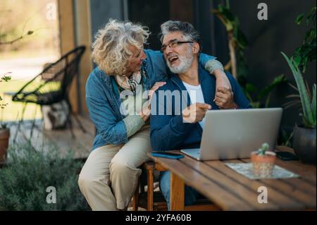 Ein Paar mittleren Alters lächelt selbstbewusst mit dem Laptop im Garten. Stockfoto