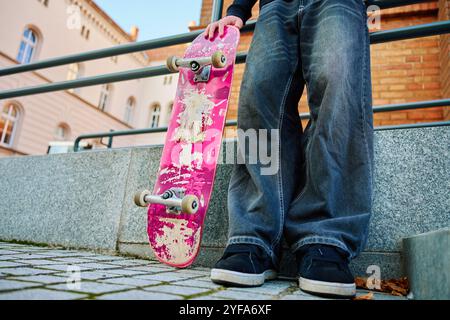 Skateboarder in Baggy-Jeans mit einem zerkratzten rosa Skateboard auf der City Street. Der Teenager ist Skateboarding. Konzept der Jugendsubkultur Stockfoto