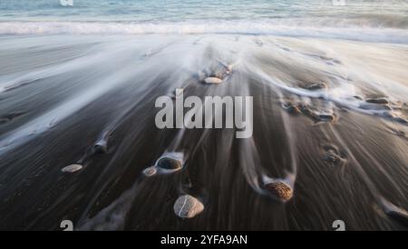 Kieselsteine am Strand auf schwarzem Sand und fließendes Meerwasser schaffen schöne Texturen. Langbelichtungsbild Stockfoto