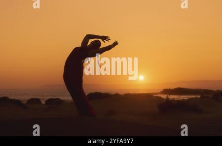 Eine Frau, die bei Sonnenuntergang Yoga am Sandstrand macht. Eine weiße Frau meditiert draußen an der Küste Stockfoto