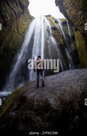 Junger Mann, der am Fuße des versteckten Wasserfalls Gljúfrabúi in Island steht, umgeben von moosbedeckten Klippen und Nebel aus dem Wasserfall Stockfoto