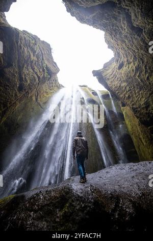 Junger Mann, der am Fuße des versteckten Wasserfalls Gljúfrabúi in Island steht, umgeben von moosbedeckten Klippen und Nebel aus dem Wasserfall Stockfoto