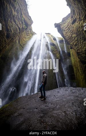 Junger Mann, der am Fuße des versteckten Wasserfalls Gljúfrabúi in Island steht, umgeben von moosbedeckten Klippen und Nebel aus dem Wasserfall Stockfoto