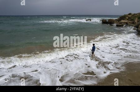 Drohnenantenne eines Jungen in Shorts, der am Sommerstrand steht und die stürmischen Wellen der spindrift betrachtet. Ansicht von hinten. Stürmisches Wetter auf der Stockfoto