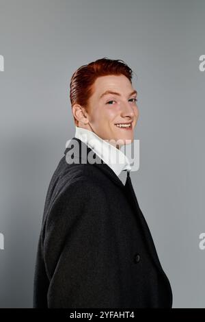 Ein selbstbewusster junger Mann mit leuchtend roten Haaren lächelt warm in einem schicken Wintermantel auf grauem Hintergrund. Stockfoto