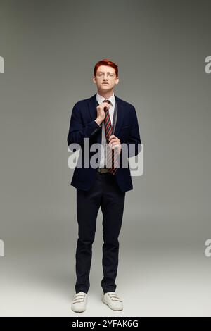 Ein stilvoller junger Mann mit rotem Haar passt seine Krawatte an und verkörpert in einem Studio raffinierte Wintermode. Stockfoto