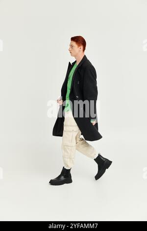 Ein junger Mann mit auffälligen roten Haaren zeigt einen modischen Wintermantel, während er in einem modernen Studio spaziert. Stockfoto