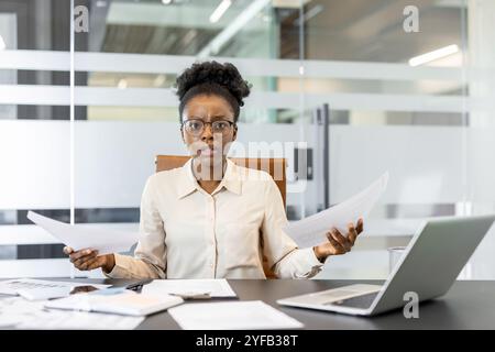 Eine junge afroamerikanische Geschäftsfrau, die Papiere hält und am Schreibtisch verwirrt aussieht. Büroumgebung mit Laptop, Dokumente deuten auf Stress und Entscheidungsfindung im geschäftlichen Umfeld hin. Stockfoto