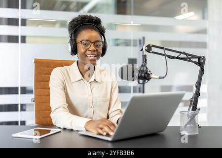Junge afroamerikanische Geschäftsfrau mit Laptop und Mikrofon, um Podcasts aufzunehmen. Faszinierende Technologie und Kopfhörer. Vertrauen in moderne Büroumgebungen, um Ideen auszutauschen. Stockfoto