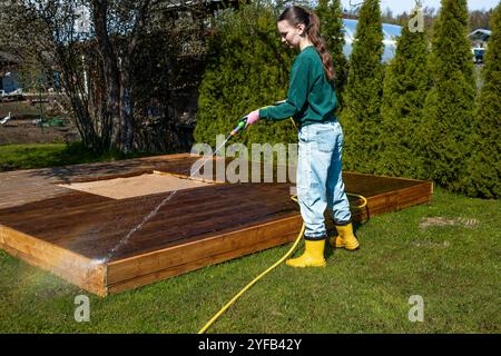 Eine junge Frau putzt eine Holzterrasse mit einem Schlauch in einem sonnigen Hinterhof Stockfoto