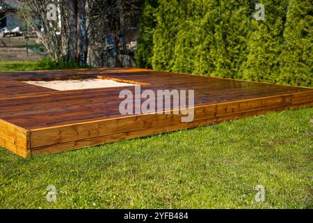 Frisch gebeizte Holzterrasse mit einem zentralen Sandkasten in einem Hinterhof umgeben von Bäumen und Büschen Stockfoto