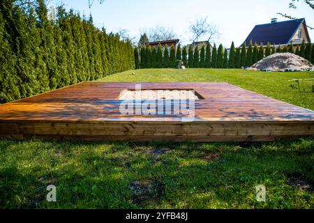 Frisch gebeizte Holzterrasse mit einem zentralen Sandkasten in einem Hinterhof umgeben von Bäumen und Büschen Stockfoto