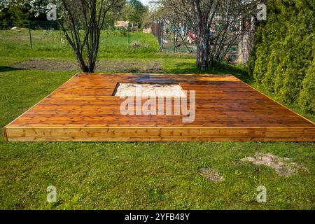 Frisch gebeizte Holzterrasse mit einem zentralen Sandkasten in einem Hinterhof umgeben von Bäumen und Büschen Stockfoto