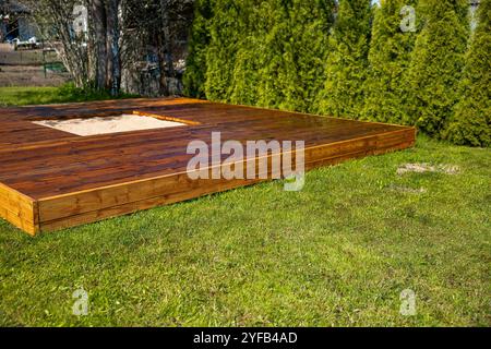 Frisch gebeizte Holzterrasse mit einem zentralen Sandkasten in einem Hinterhof umgeben von Bäumen und Büschen Stockfoto