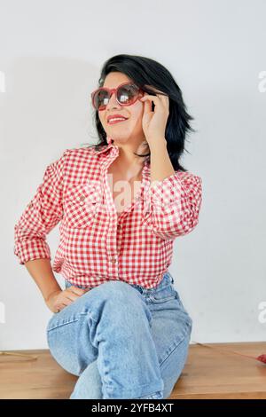 latina Frau mit schwarzen Haaren und roten Lippen mit rotem kariertem Hemd und Jeans, roter Sonnenbrille mit positiver und fröhlicher Einstellung und weißem Hintergrund Stockfoto