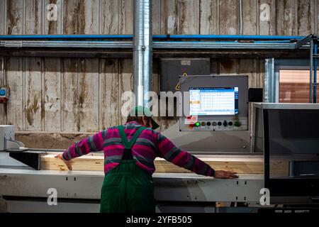 Ein Arbeiter bedient eine Maschine in einer Holzbearbeitungsfabrik, die Holzdielen verarbeitet, im Hintergrund Holzstapel und Industrieanlagen Stockfoto