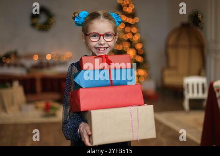 Ein fröhliches kleines Mädchen in stilvoller Brille und einem wunderschönen festlichen Kleid steht neben einem glitzernden Weihnachtsbaum und legt ein wunderschön gewickeltes Geschenk in ihre Hände. Moment, um ihr Geschenk auszupacken, das den zauberhaften Geist des Weihnachtsstils verkörpert. Stockfoto