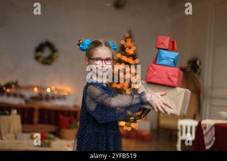 Ein fröhliches kleines Mädchen in stilvoller Brille und einem wunderschönen festlichen Kleid steht neben einem glitzernden Weihnachtsbaum und legt ein wunderschön gewickeltes Geschenk in ihre Hände. Moment, um ihr Geschenk auszupacken, das den zauberhaften Geist des Weihnachtsstils verkörpert. Stockfoto