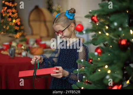 Ein fröhliches kleines Mädchen in stilvoller Brille und einem wunderschönen festlichen Kleid steht neben einem glitzernden Weihnachtsbaum und legt ein wunderschön gewickeltes Geschenk in ihre Hände. Moment, um ihr Geschenk auszupacken, das den zauberhaften Geist des Weihnachtsstils verkörpert. Stockfoto